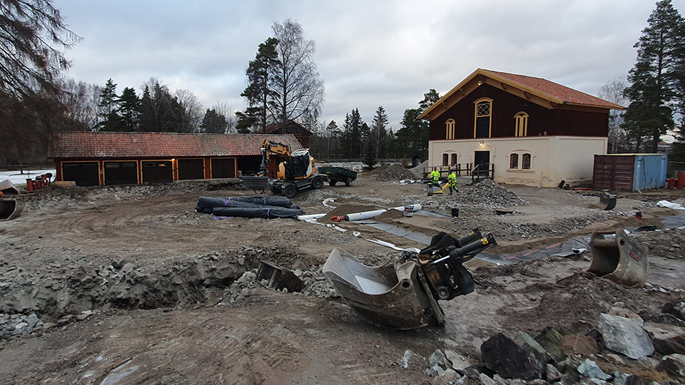Bild över centrala gruvområdet vid Sala Silvergruva. På området syns två män i gula skyddskläder som arbetar med åtgärderna, området har schaktats och rullar med bentonitmattor syns, i bakgrunden syns gruvstallet 
